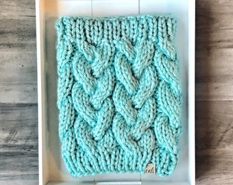 May include: A turquoise cable-knit cowl neck warmer. The chunky knit has a textured, braided design. The cowl is displayed in a white tray on a wooden surface. A small tag with the word "cali" is attached to the cowl.
