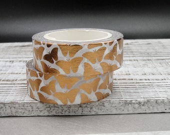 May include: Two rolls of decorative washi tape. The tape features a repeating pattern of gold butterflies on a white background. The rolls are stacked on top of each other, showcasing the design. The tape is ideal for crafts and scrapbooking.