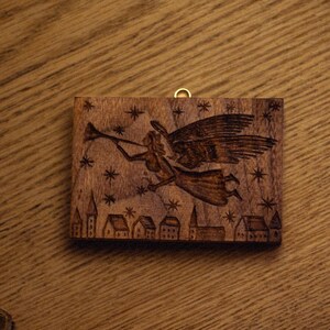 May include: A wooden wall hanging with a carved design of an angel playing a trumpet, stars, and a village. The artwork is rectangular with a gold-colored loop for hanging. The piece is set on a wooden surface.