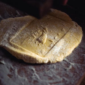 May include: A close-up shot of a piece of dough with a detailed embossed design. The dough is dusted with flour and sits on a dark wooden surface. The design appears to be a religious icon.