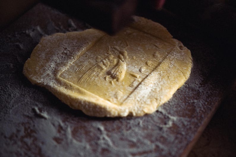 May include: A close-up shot of a piece of dough with a detailed embossed design. The dough is dusted with flour and sits on a dark wooden surface. The design appears to be a religious icon.