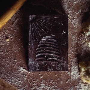 May include: A dark brown, rectangular wooden stamp with a detailed relief of a beehive, sun rays, and flying bees. The stamp is surrounded by flour and a rolling pin, suggesting a baking theme.