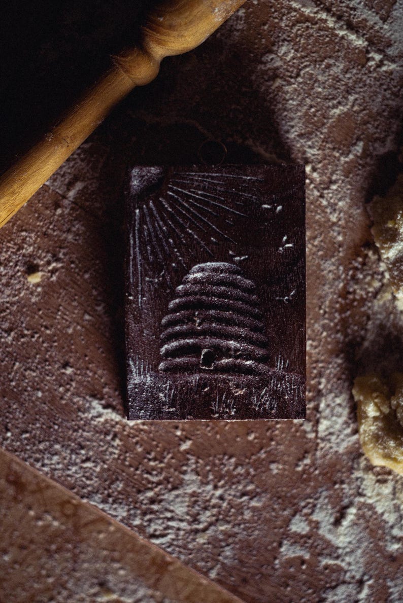 May include: A dark brown, rectangular wooden stamp with a detailed relief of a beehive, sun rays, and flying bees. The stamp is surrounded by flour and a rolling pin, suggesting a baking theme.