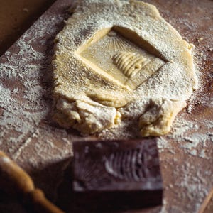 May include: A wooden cutting board with a rectangular piece of dough, dusted with flour, featuring a carved design. A wooden rolling pin and a carved wooden block are also present, suggesting baking or food preparation.