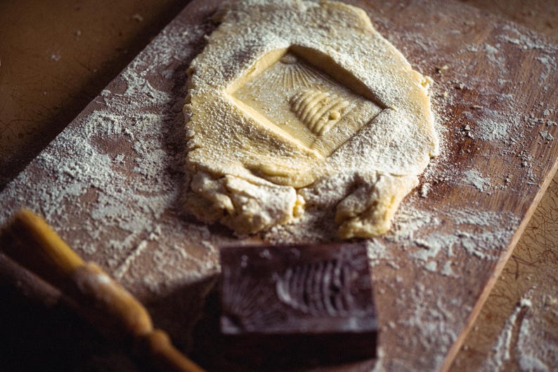 May include: A wooden cutting board with a rectangular piece of dough, dusted with flour, featuring a carved design. A wooden rolling pin and a carved wooden block are also present, suggesting baking or food preparation.