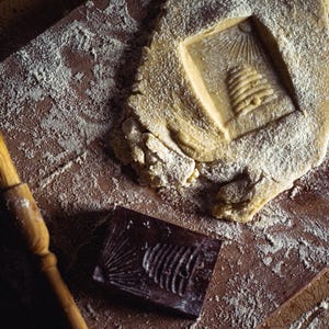 May include: A wooden pastry board with flour, a piece of dough with a beehive imprint, a wooden brush, and a dark brown mold with a fish design. The scene suggests baking or crafting, with a rustic, artisanal feel.