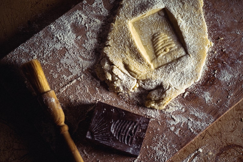 May include: A wooden pastry board with flour, a piece of dough with a beehive imprint, a wooden brush, and a dark brown mold with a fish design. The scene suggests baking or crafting, with a rustic, artisanal feel.