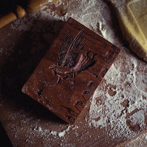 May include: A dark brown wooden cookie mold with a carved design of a bird with outstretched wings, surrounded by stars. The mold is rectangular and rests on a wooden surface dusted with flour, alongside a rolling pin and a piece of dough.
