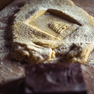May include: Close-up of a wooden surface with a rectangular dough piece dusted with flour. The dough has a carved square with a beehive design. A dark brown rectangular object is in the foreground.
