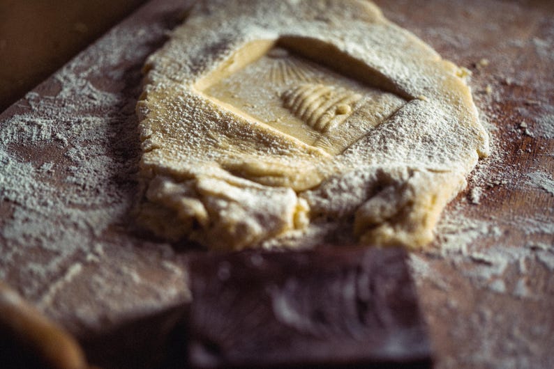 May include: Close-up of a wooden surface with a rectangular dough piece dusted with flour. The dough has a carved square with a beehive design. A dark brown rectangular object is in the foreground.
