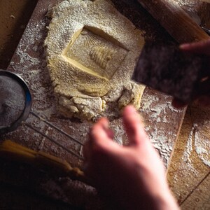 May include: A close-up shot of a baking scene. A square-shaped cookie dough is stamped with a design. A small sieve, rolling pin, and pastry brush are also visible. The scene is dusted with flour, suggesting a home baking activity.
