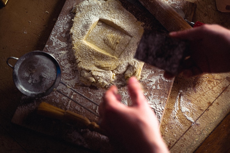 May include: A close-up shot of a baking scene. A square-shaped cookie dough is stamped with a design. A small sieve, rolling pin, and pastry brush are also visible. The scene is dusted with flour, suggesting a home baking activity.