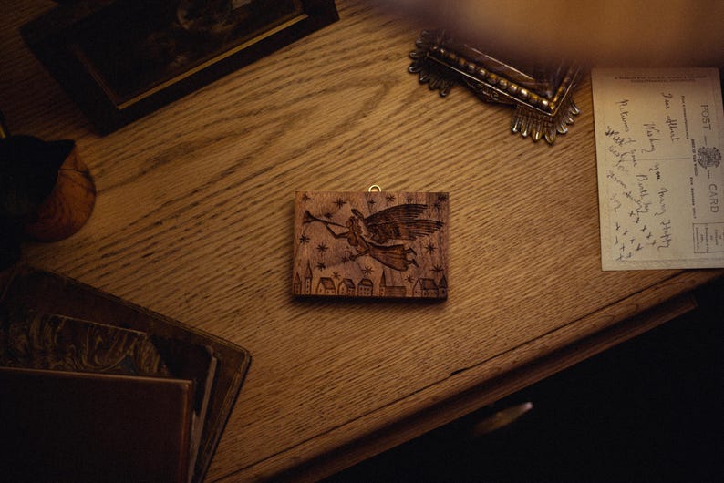 May include: A wooden wall hanging with a carved design of an angel playing a trumpet, stars, and a village. The artwork is rectangular with a gold-colored loop for hanging. The piece is set on a wooden surface.