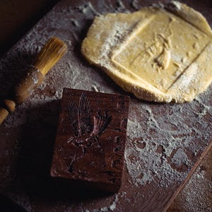 May include: A wooden cookie mold with a carved bird design, a pastry brush, and a piece of dough with an imprinted bird. The items are on a wooden surface dusted with flour, suggesting baking.