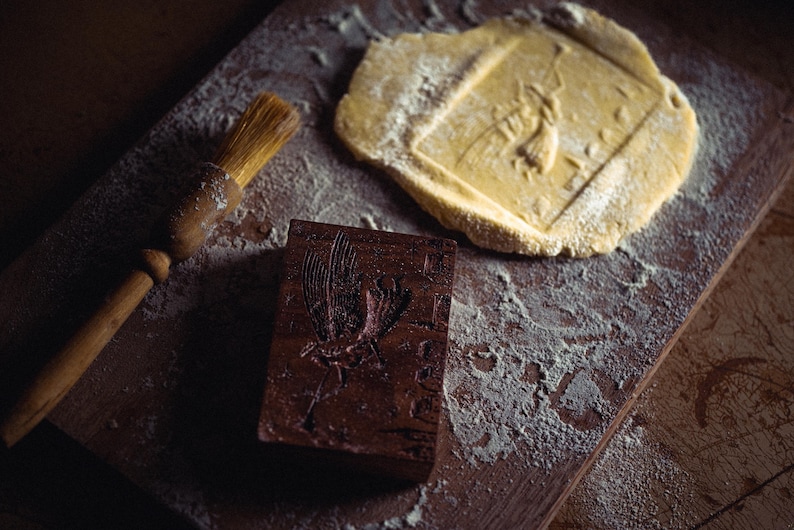 May include: A wooden cookie mold with a carved bird design, a pastry brush, and a piece of dough with an imprinted bird. The items are on a wooden surface dusted with flour, suggesting baking.