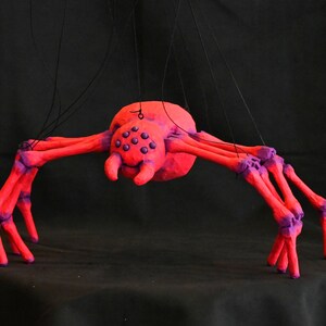 May include: A red and purple spider puppet with eight legs and black strings. The spider has a large, round head with purple eyes.