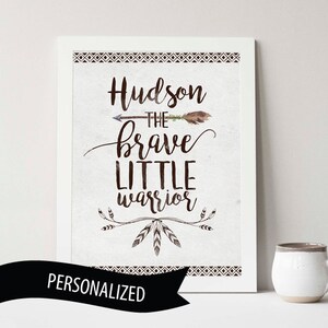 May include: A white framed print with a brown background and the text "Hudson the brave little warrior" in brown cursive font. The text is surrounded by a brown arrow, feathers, and a tribal pattern. The bottom of the print has a black ribbon with the word "PERSONALIZED" in white text.