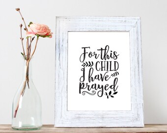 Puede incluir: Una impresión enmarcada en blanco con texto negro que dice "For this child I have prayed". La impresión está sobre una superficie de madera con un jarrón de flores rosas en el fondo.
