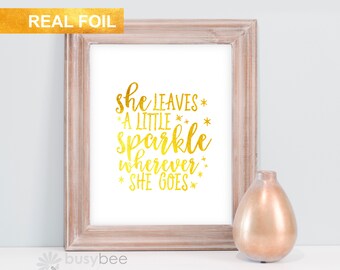 Puede incluir: Una impresión enmarcada con el texto "she leaves a little sparkle wherever she goes" en letras doradas. La impresión está en un marco de madera marrón claro, y hay un jarrón decorativo al lado.