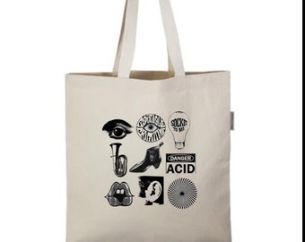 May include: A cream-coloured tote bag featuring black graphic illustrations such as an eye, lips, a boot, and the word "ACID" in a warning sign. The bag has long, matching handles.