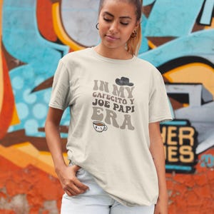 May include: A cream-colored t-shirt with the text "IN MY CAFECITO Y JOE PAPI ERA" in brown and gray lettering. The shirt features a small cowboy hat graphic and a coffee cup illustration. The model is wearing white shorts.
