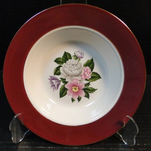 May include: A white ceramic soup bowl with a burgundy rim and a gold accent. The bowl features a floral design with pink, white, and purple flowers.