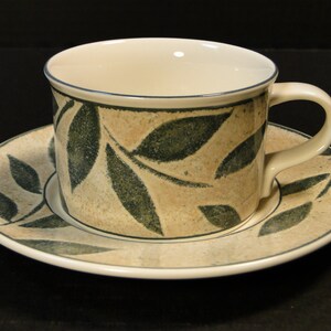 May include: A cream-colored teacup and saucer with a green leaf pattern. The cup has a handle and the saucer is round.