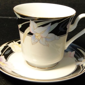 May include: A white teacup and saucer with a black and gold floral design. The flowers are blue, pink, and yellow.