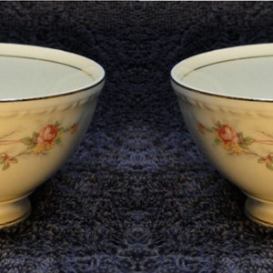 May include: Two cream-colored teacups with floral designs. Each cup features a handle, a footed base, and a gold trim along the rim and base. The floral pattern includes pink and yellow roses with green leaves.