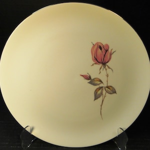 May include: A white ceramic dinner plate with a gold rim features a single pink rose with green leaves and stems.
