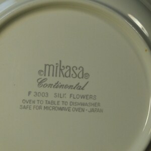 Pu&ograve; includere: Primo piano di un piatto bianco Mikasa Continental. Il piatto presenta il testo "mikasa Continental F 3003 SILK FLOWERS OVEN TO TABLE TO DISHWASHER SAFE FOR MICROWAVE OVEN JAPAN". Il piatto &egrave; di colore bianco sporco.