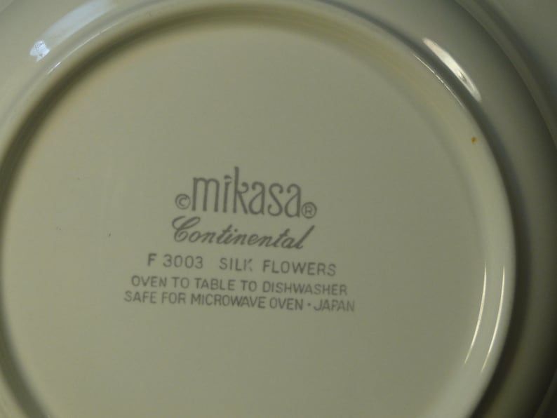 Pu&ograve; includere: Primo piano di un piatto bianco Mikasa Continental. Il piatto presenta il testo "mikasa Continental F 3003 SILK FLOWERS OVEN TO TABLE TO DISHWASHER SAFE FOR MICROWAVE OVEN JAPAN". Il piatto &egrave; di colore bianco sporco.