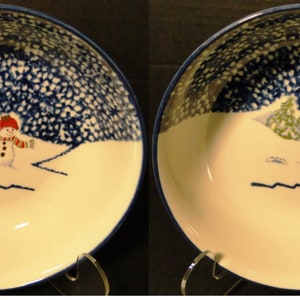 May include: A ceramic bowl featuring a hand-painted winter scene with a snowman wearing a red hat and a striped scarf. The background includes a dark blue textured pattern and a green pine tree. The bowl has a smooth cream-colored interior.