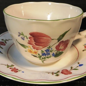 May include: A white teacup and saucer with a green rim and floral pattern. The pattern features pink, red, yellow, blue, and green flowers.