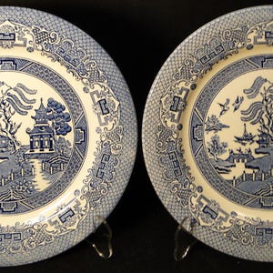 May include: Two blue and white ceramic dinner plates with a willow pattern design. The plates have a white background with a blue border and a blue willow tree design in the center.