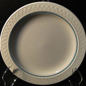 May include: A round off-white ceramic plate featuring a textured lattice pattern embossed along the wide rim. A thin, solid blue circular band is painted on the inner edge of the rim, encircling the smooth, plain center of the plate.
