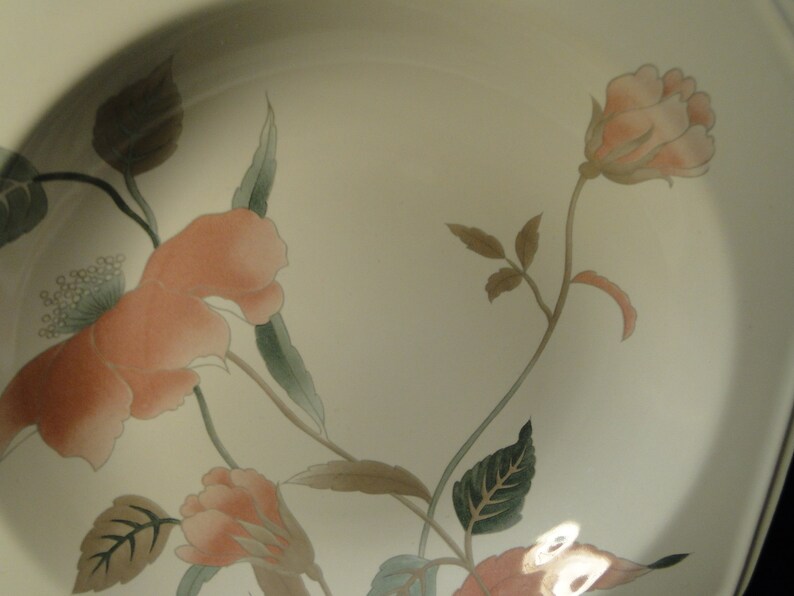 Pu&ograve; includere: Piatto in ceramica color crema con un motivo floreale. Il design presenta rose color pesca, foglie verdi e steli marroni. Il piatto ha un bordo leggermente curvo e una superficie liscia.