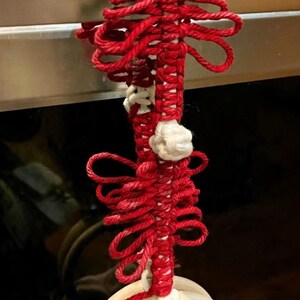 May include: A handmade red and white decorative knot, possibly a towel holder. The knot is made of red rope with white accents and a wooden ring at the bottom. The knot is hanging from a metal surface.