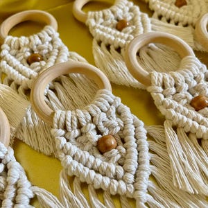 Op de afbeelding: Close-up van verschillende crèmekleurige macrame onderzetters met houten ringen en kralen. De onderzetters hebben een ruitpatroon en franjes aan de onderkant. De achtergrond is goudgeel.