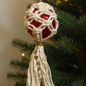May include: A handmade Christmas ornament featuring a red glitter ball encased in a cream-colored macrame net. The ornament has a cream tassel with small gold bells and is hanging from a Christmas tree.
