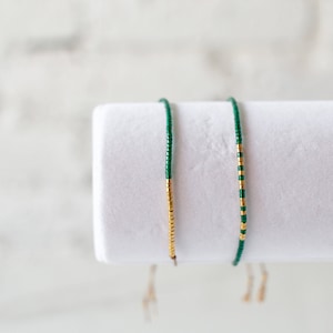May include: Two beaded bracelets on a white display stand. One bracelet is green with gold accents, and the other is gold with green accents.