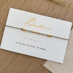 Op de afbeelding: Een minimalistische armband met kleine gouden en lichtblauwe kralen geregen op een bruin koord. De armband ligt op een witte kaart met het woord "Badass" in gouden script en de tekst "Be Strong. Be Brave. Be Badass."