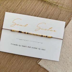 Puede incluir: Una pulsera de amistad con cuentas doradas y marrones se muestra en una tarjeta blanca. La tarjeta dice "Soul Sister" en letras doradas, con el texto "Friends by heart. And sister by soul."