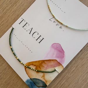 May include: Two beaded necklaces, one light green and one dark green, are displayed on a card with the word "TEACH" printed in black. The necklaces have gold-colored beads. The card also has the words "Live, Love" printed on it.