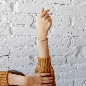 May include: A person's arm with a stack of gold and white beaded bracelets on their wrist. The arm is raised and the hand is pointing upwards. The person is wearing a brown sweater.