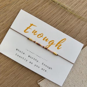 Op de afbeelding: Een armband met roze en gouden kralen wordt getoond op een witte kaart. De kaart heeft het woord "Enough" in oranje script, samen met de woorden "Whole. Worthy. Enough. Exactly as you are."