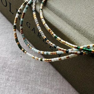 Puede incluir: Un collar de cuentas multicadena con cuentas doradas, verdes y blancas. El collar está en capas y tiene un borla al final.