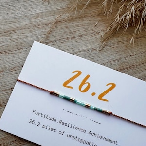 Puede incluir: Un brazalete de cuerda marrón con cuentas turquesa y doradas. El brazalete tiene el número "26.2" escrito en una tarjeta blanca. La tarjeta también dice "Fortitude. Resilience. Achievement. 26.2 miles of unstoppable strength."