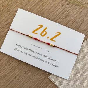 May include: A white card with the number "26.2" in gold lettering, with a red string bracelet. The card also has the text "Fortitude. Resilience. Achievement. 26.2 miles of unstoppable strength."