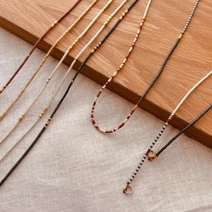Puede incluir: Una colección de delicados collares de cuentas en varios colores, incluyendo marrón, crema, negro y rojo. Cada collar presenta pequeñas cuentas coloridas y detalles dorados. Los collares se exhiben sobre una superficie de madera.